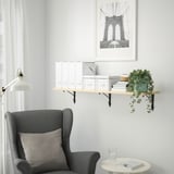 Minimalist reading nook: gray chair, floating shelf with white decor, hanging plant, brooklyn bridge art.