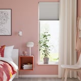 Cozy bedroom with floral bedding, pink walls, TREDANSEN blind, plant, and side table with lamp and books.