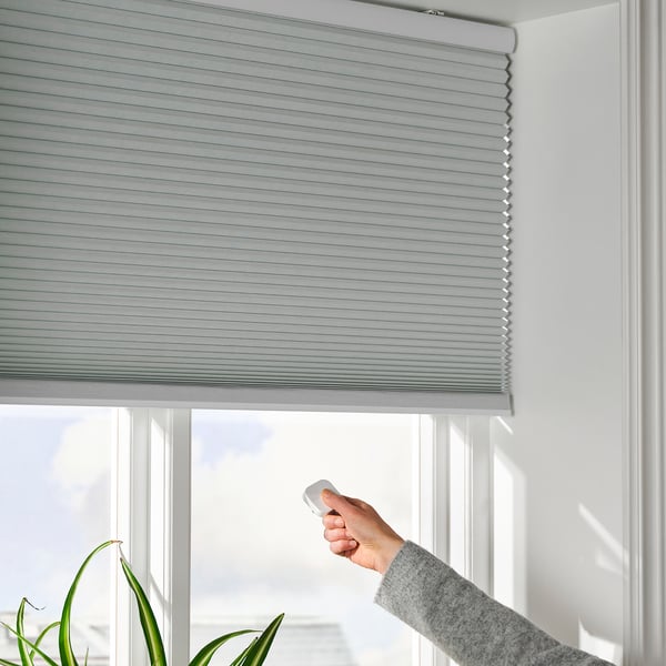Hand raises remote to control motorized blinds over window.
