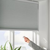 Hand raises remote to control motorized blinds over window.