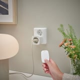 Hand holding remote controls smart plug; connected lamp, flowers, wall socket.