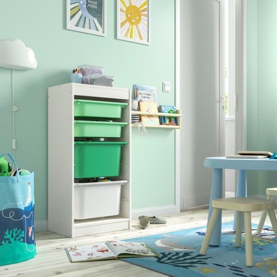 A TROFAST storage unit in white with green bins, has open shelves and is placed in a brightly lit room, next to a small table and chair set.