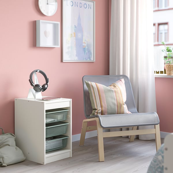 Small white TROFAST storage unit beside a grey chair with a striped pillow. Headphones are on top. Light wood floor.