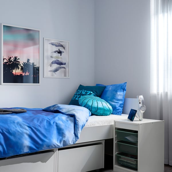 Bedroom with blue bedding and pillows, matching wall art, featuring a white TROFAST storage unit beside it.