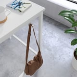 Brown leather bag hangs on white metal table leg. Table has white cup and book. Plant beside.
