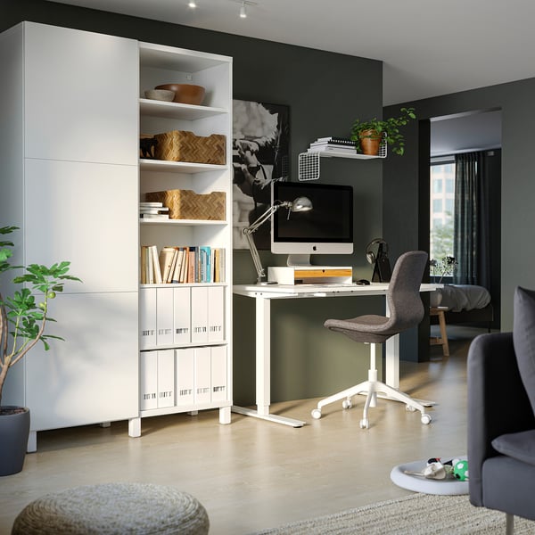 Modern home office with white TROTTEN desk, grey chair, and green walls. Shelves hold books and decor. Computer, lamp, and plant. Light-colored floor.