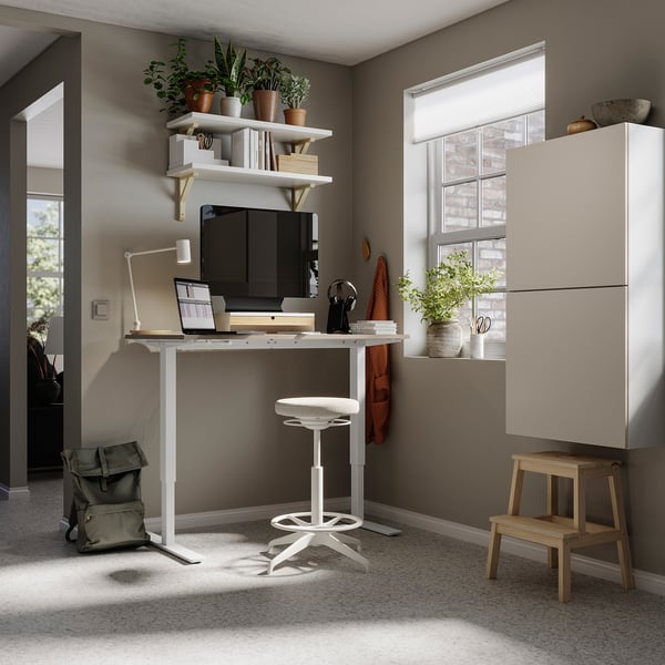 Home office with white TROTTEN desk, stool, shelves, and cabinets. Plants, laptop, and monitor on desk. Natural light from window.