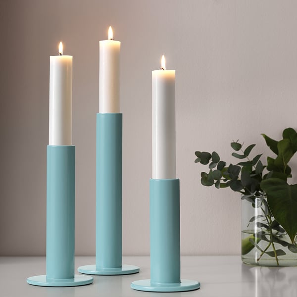Three blue ceramic candle holders of varying heights with lit white candles on a table, accompanied by a vase of greenery.
