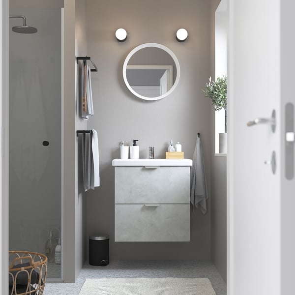 Minimalist bathroom: round mirror, concrete vanity, glass shower, wall towels, plant.