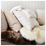 Image soft sheepskin rug, 3 square pillows on it. Sheepskin: off-white, thick, fluffy; pillows: beige, light gray, white.