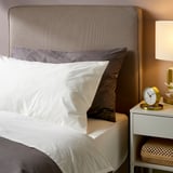 Bedroom with ULLVIDE gray pillowcase on white bed. Neutral colors, smooth sheets, nicely arranged bedside with clock and lamp.