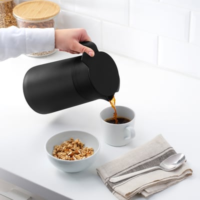Person pours coffee from flask into mug next to granola, spoon, and napkins.