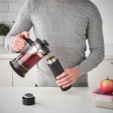 Pouring coffee into black travel mug on kitchen counter.