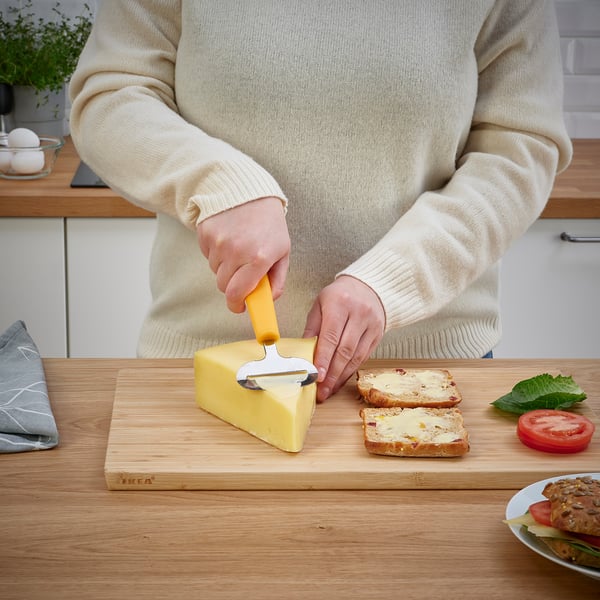 Person slicing cheese with a yellow UPPFYLLD cheese slicer on a wooden cutting board.