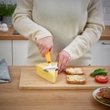 Person slicing cheese with a yellow UPPFYLLD cheese slicer on a wooden cutting board.