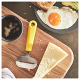 Bright yellow cheese slicer lies on wooden board with cheese and coffee nearby.