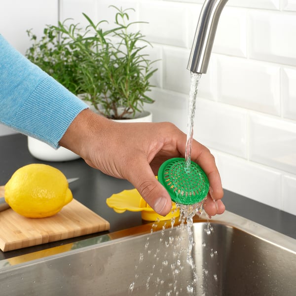 Person washing a green lemon squeezer under a tap, with lemon and herb background.