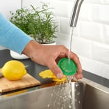 Person washing a green lemon squeezer under a tap, with lemon and herb background.