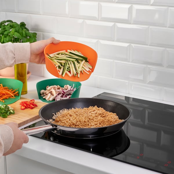 A person cooks in the kitchen with sliced cucumbers and noodles. Chopped vegetables and a cutting board are nearby.