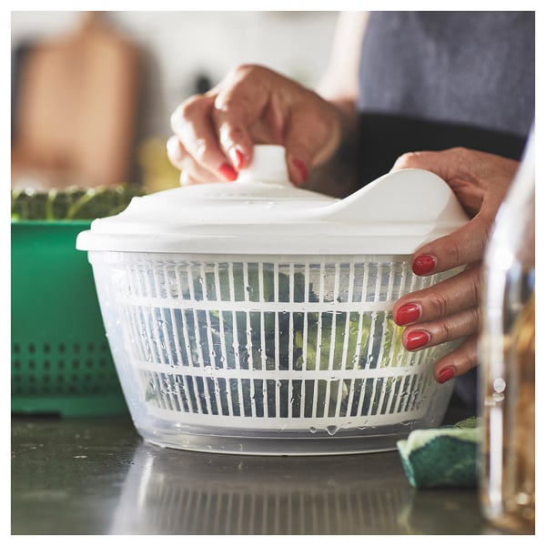 Person uses white UPPFYLLD salad spinner, rinsing greens in kitchen. Plastic, round, with handle, sits on counter.