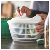 Person uses white UPPFYLLD salad spinner, rinsing greens in kitchen. Plastic, round, with handle, sits on counter.