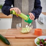 Person uses green UPPFYLLD vegetable slicer to cut cucumber into strips over bowl on wooden board.