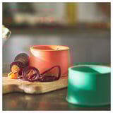 Two colorful vegetable slicers, orange and green, sit on a wooden surface. Orange slicer has a handle and blade, designed for long veg strips.