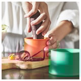 Person using orange UPPFYLLD vegetable slicer on wooden board with green container nearby, peeling red vegetable.