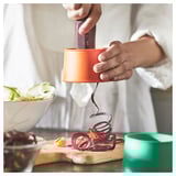 Person using orange UPPFYLLD vegetable slicer to cut red vegetable into spiral strips on wooden board.