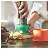 A person peels a cucumber using a green UPPFYLLD vegetable slicer on a wooden board with orange slicer beside.