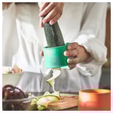 Person peels cucumber using green UPPFYLLD vegetable slicer, creating long strips on wooden board.