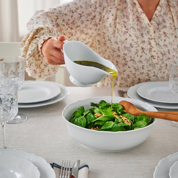 A person pours dressing from a white gravy boat onto a salad in a white bowl on a table set with plates and glasses.
