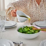 A person pours dressing from a white gravy boat onto a salad in a white bowl on a table set with plates and glasses.