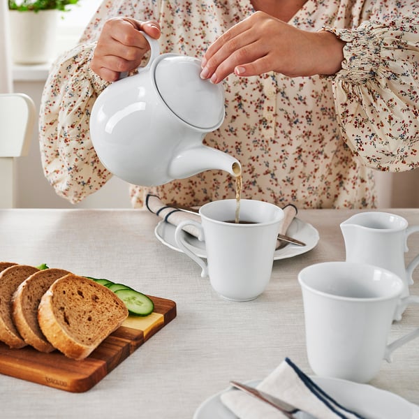 Person pouring tea into white mug, with sliced bread and cucumber on table.