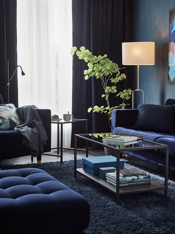 Modern living room with dark blue furniture, silver lamp, glass table, and blue rug.
