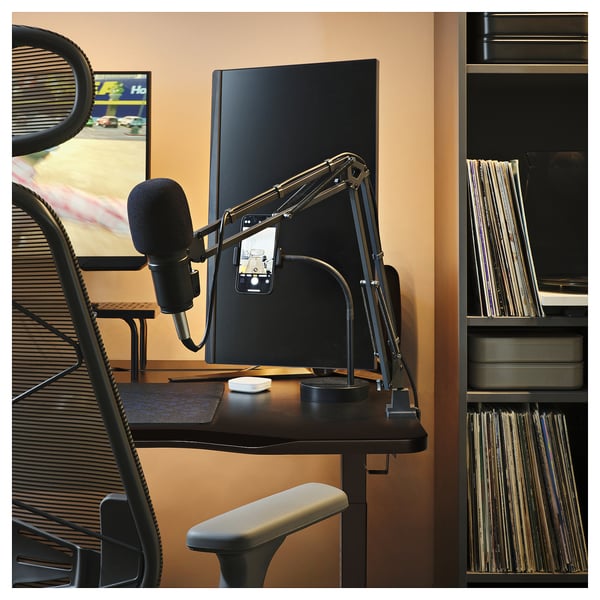 A black gaming desk with a high-tech setup. A monitor, phone with mic, and vinyl records are visible.
