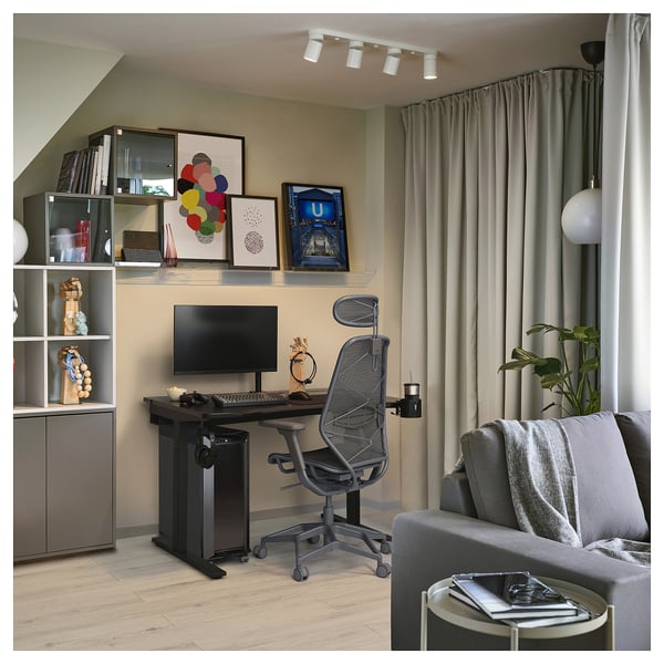 Modern home office with utmaring black desk, ergonomic chair, monitor, shelves with colorful art, gray curtains, and cozy gray sofa.