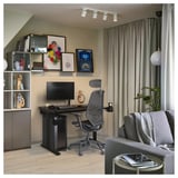 Modern home office with utmaring black desk, ergonomic chair, monitor, shelves with colorful art, gray curtains, and cozy gray sofa.