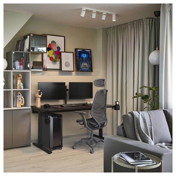 Home office setup: black desk with dual monitors, ergonomic chair, gray sofa, bookshelf with accessories, and large windows with curtains.