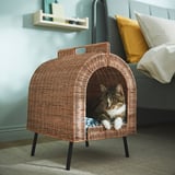 Cat inside wicker house, cushioned, elevated on legs for airflow, handle for easy transport, placed near bed.