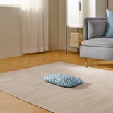 A cozy pet cushion on a rug near a grey armchair, perfect for cats or small dogs to relax and sleep.