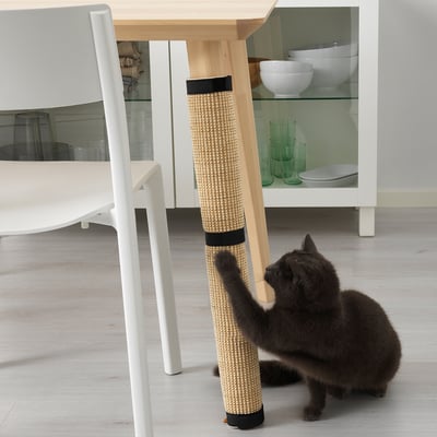 Black cat plays with beige and black UTSÅDD cat scratching mat wrapped around table leg in modern kitchen.