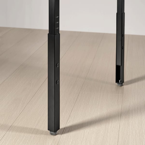 Black adjustable desk leg, sturdy with height markings, on wooden floor.