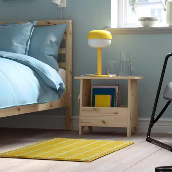 Yellow checked VÄNDPLATS rug beside bed with lamp, compact.