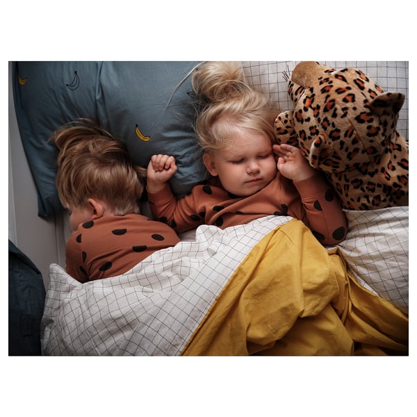 Two children sleeping, one holding a leopard pillow, under a yellow blanket, wearing matching brown pajamas with dark spots.