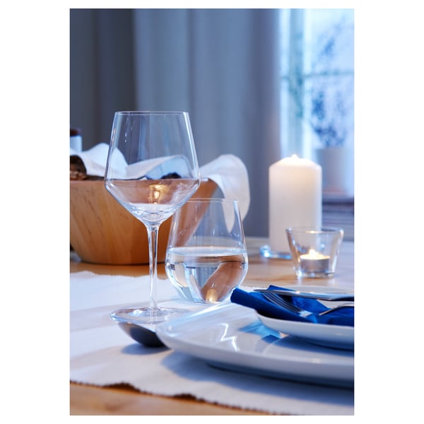 A VÄRDERA table setting with clear wine glasses, white plates, and blue napkins on a wooden table. Candles are lit in small glass holders.