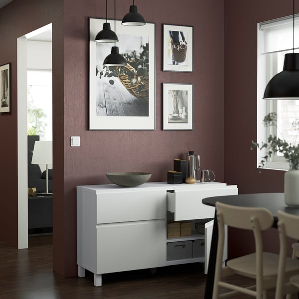 Modern dining area with white storage unit featuring integrated handles and open drawer. Decorative bowl, cups, and art on dark purple wall.