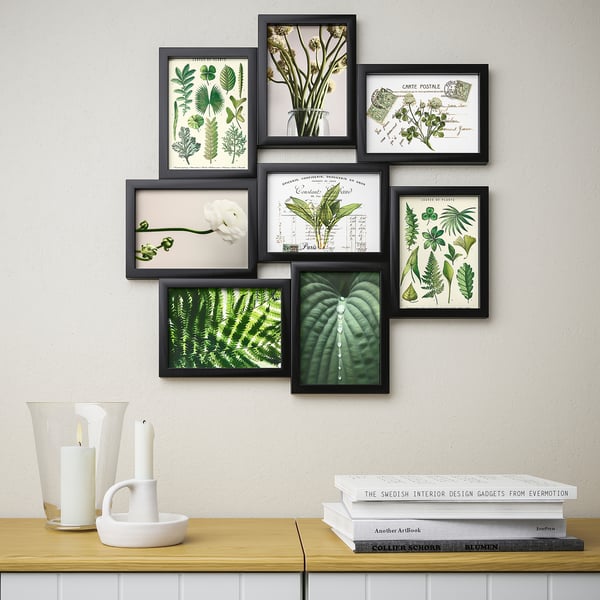 Wall-mounted collage of eight framed green plant illustrations, part of ikeas VÄXBO series, arranged above a shelf holding candles and books.
