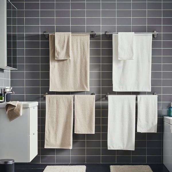 Modern bathroom with dark gray tiled walls. Four towels hang neatly in rows, two beige and two white, on silver towel rails.