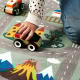 Childs hand plays with wooden car on colorful rug, showcasing its non-slip feature.